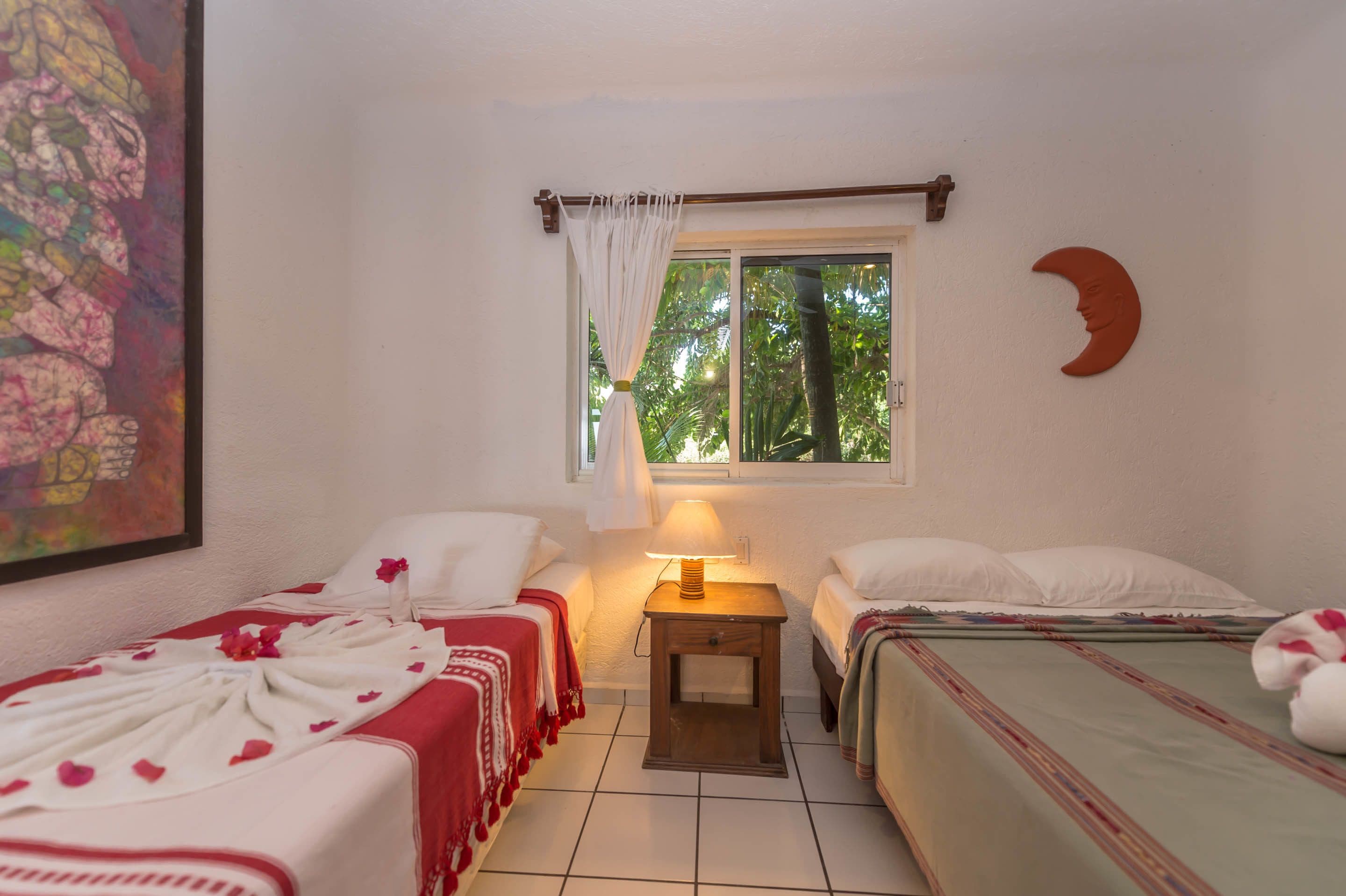 Una habitación con dos camas, una ventana con cortina, una mesita de noche con lámpara y un cuadro en la pared.