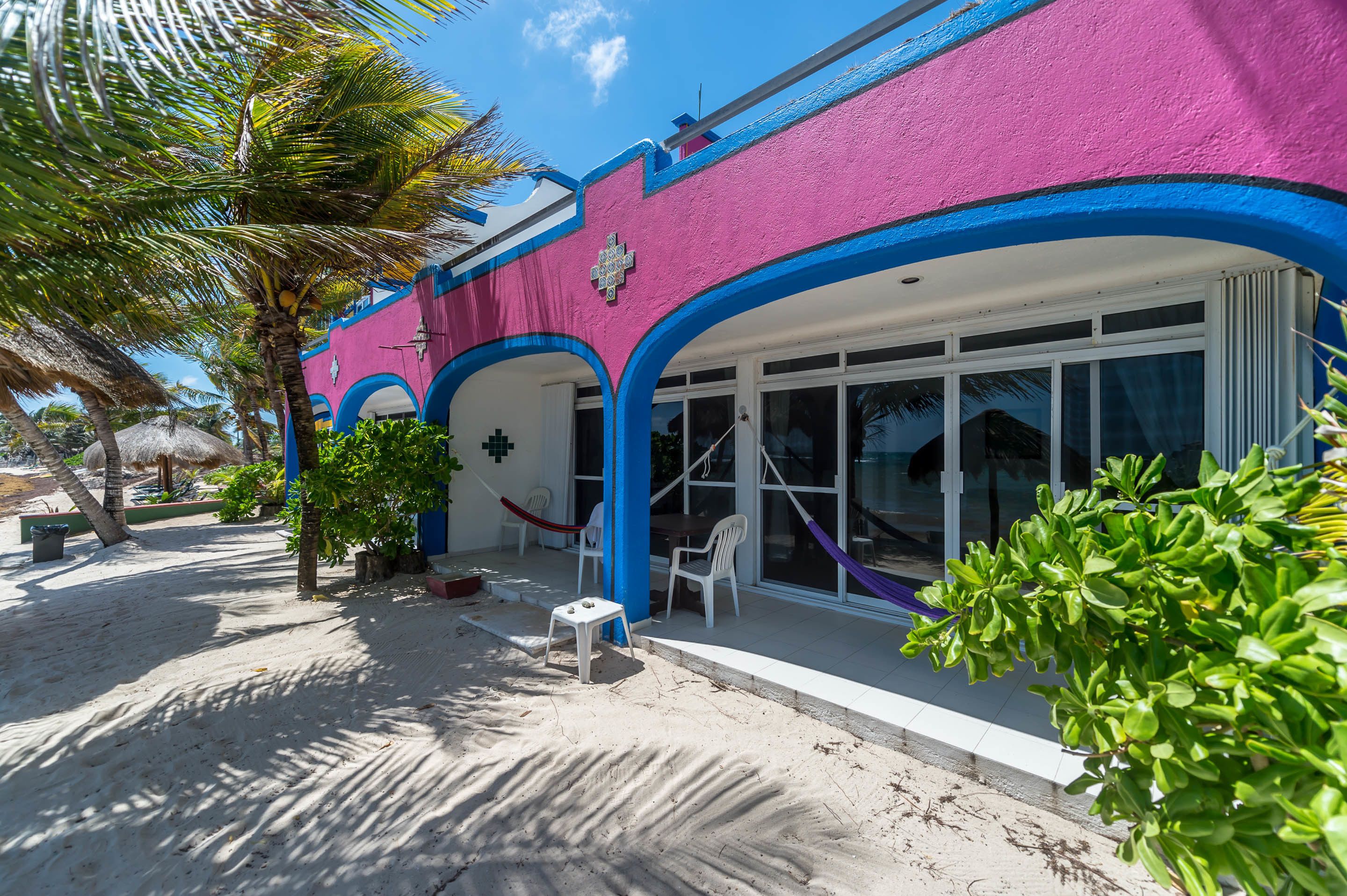 A beachside building with a vibrant pink and blue facade, large windows, and a small patio area with a table and chairs.