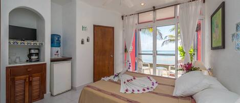 A hotel room with a bed, a small kitchenette, a ceiling fan, and a view of the ocean.