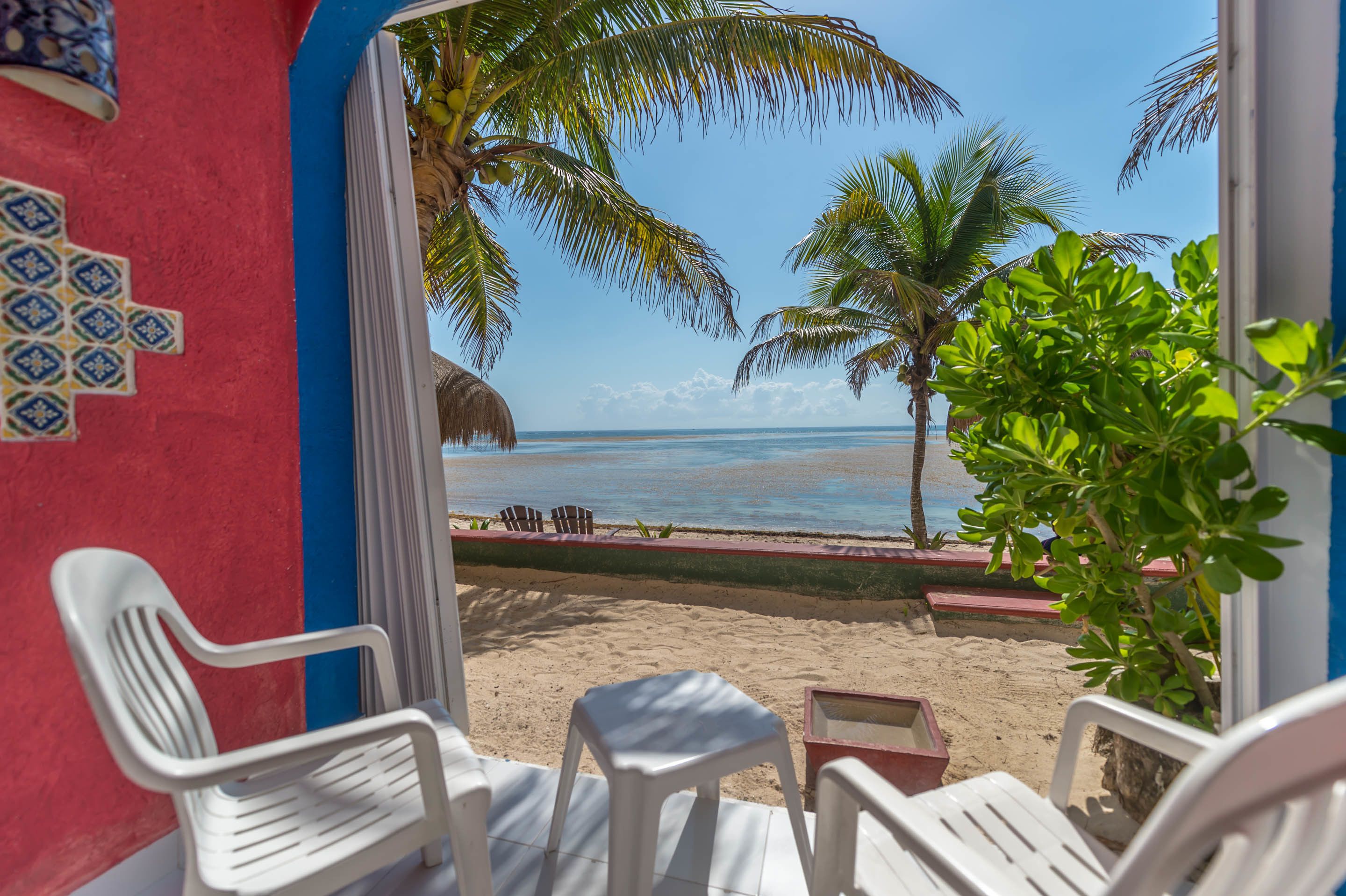 Une vue sur la plage depuis une chambre avec des chaises en plastique blanches et une petite table, un palmier et une structure dotée d’un toit de chaume.