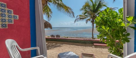 Vista a la playa desde una habitación con sillas de plástico blanco y una pequeña mesa, una palmera y una estructura de techo de paja.