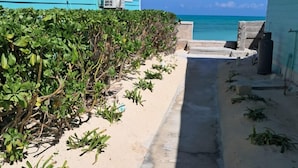 Ein schmaler Betonweg, gesäumt von einem blauen Gebäude und einer Hecke, führt zu einem Sandstrand mit Blick auf den Ozean.