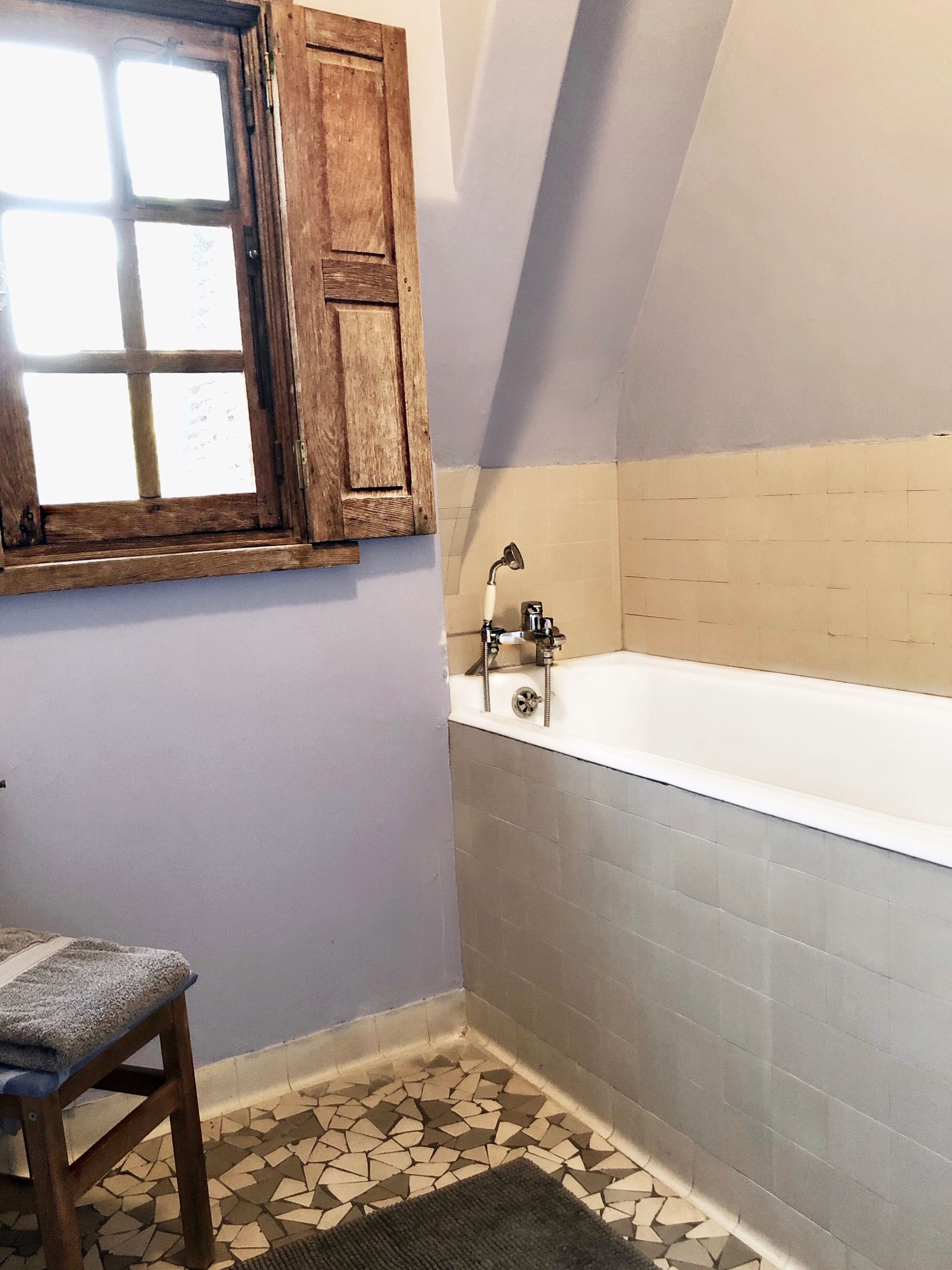 A bathroom with a bathtub, a wooden stool, a window with wooden shutters, and a tiled floor.