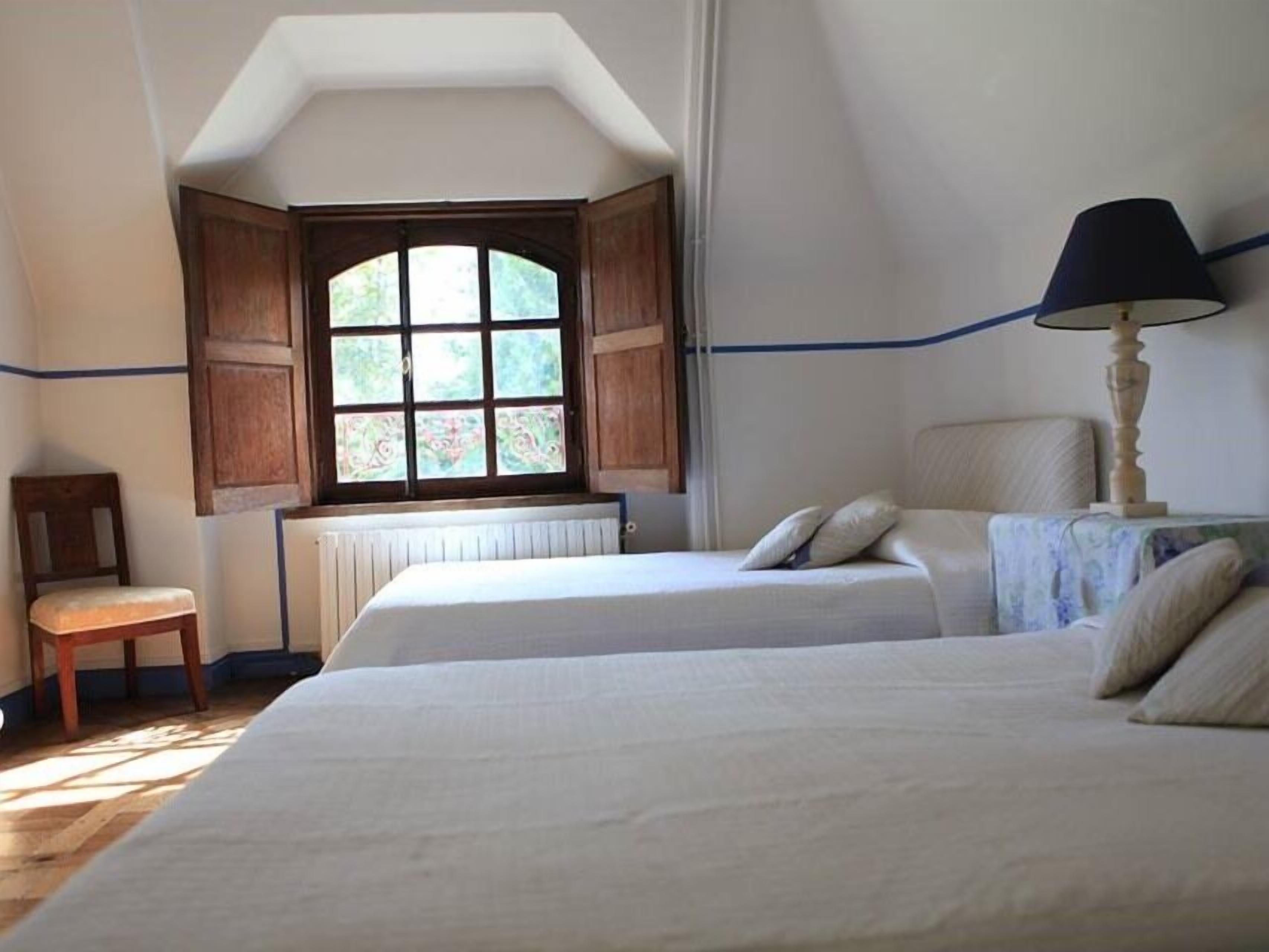 A room with two beds, a window with wooden shutters, a chair, and a lamp.
