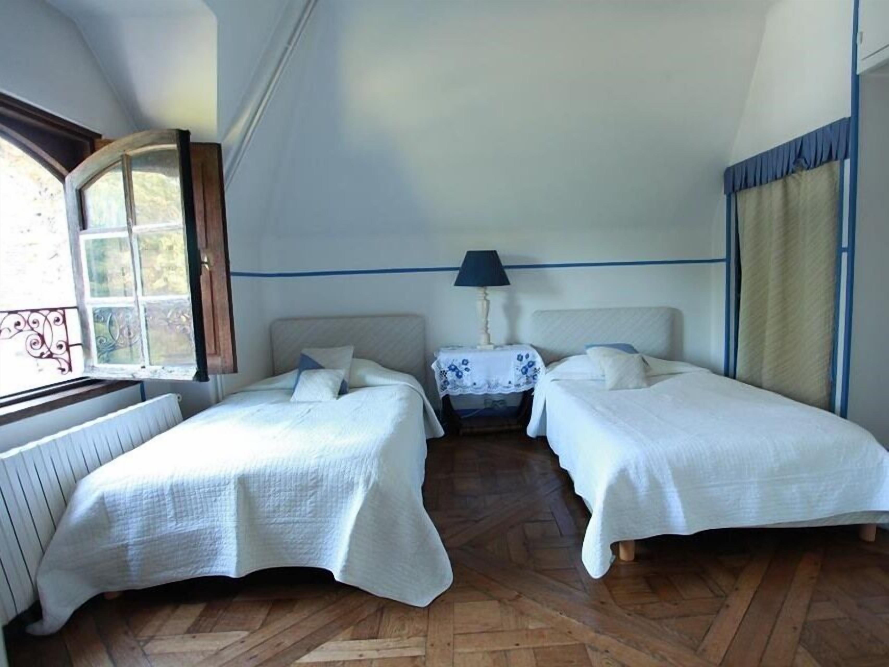 Quarto com duas camas, uma mesa de cabeceira com abajur, janela com cortinas e radiador.