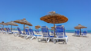 Ein Strand mit Reihen aus blauen und weißen Liegestühlen und reetgedeckten Sonnenschirmen.