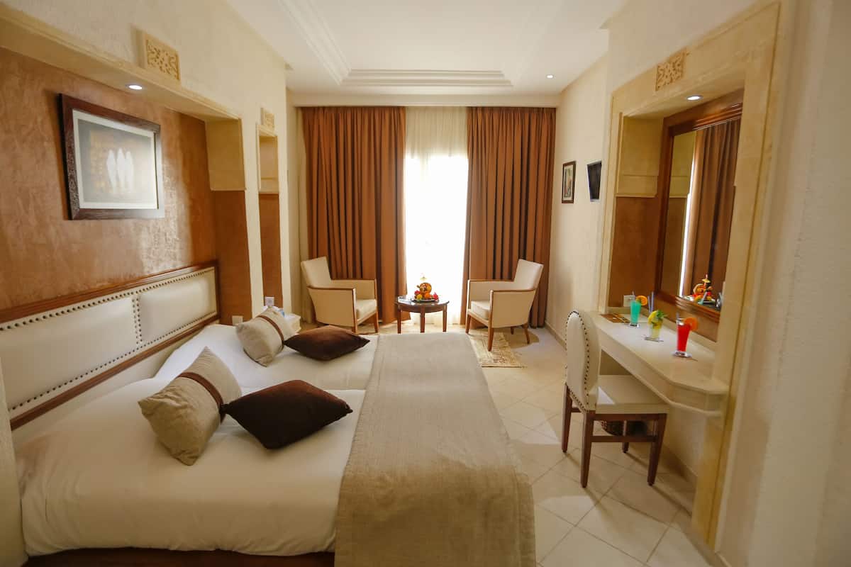 Une chambre d’hôtel avec un lit, deux chaises, un bureau avec un miroir et une petite table avec des boissons.