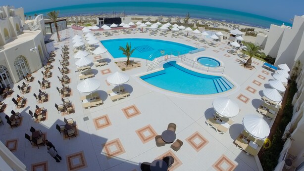 Ein Poolbereich mit mehreren Liegestühlen und Sonnenschirmen, einem zentralen Pool und Blick auf den Strand.