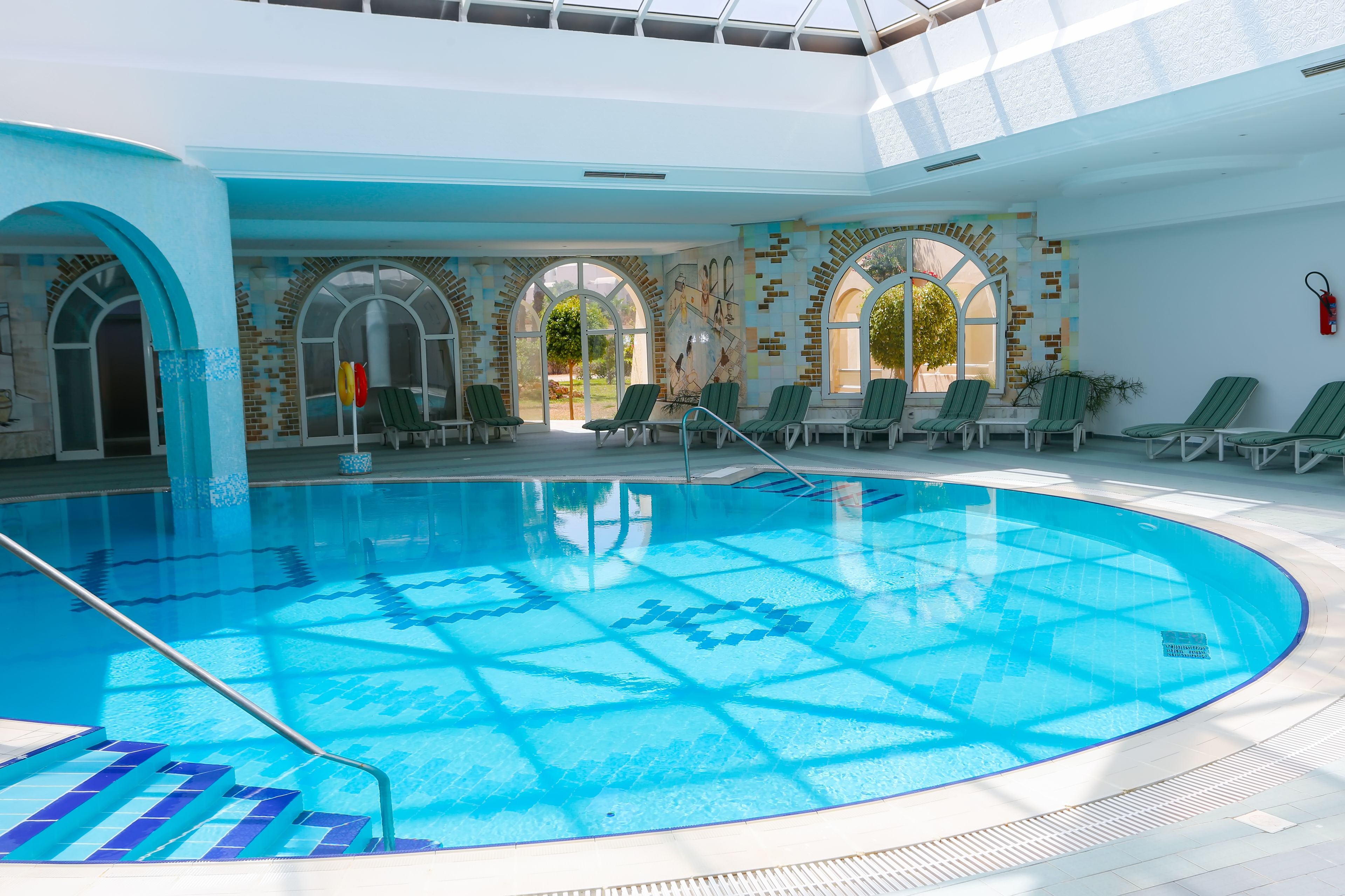 An indoor swimming pool with clear blue water, surrounded by green lounge chairs and arched windows.