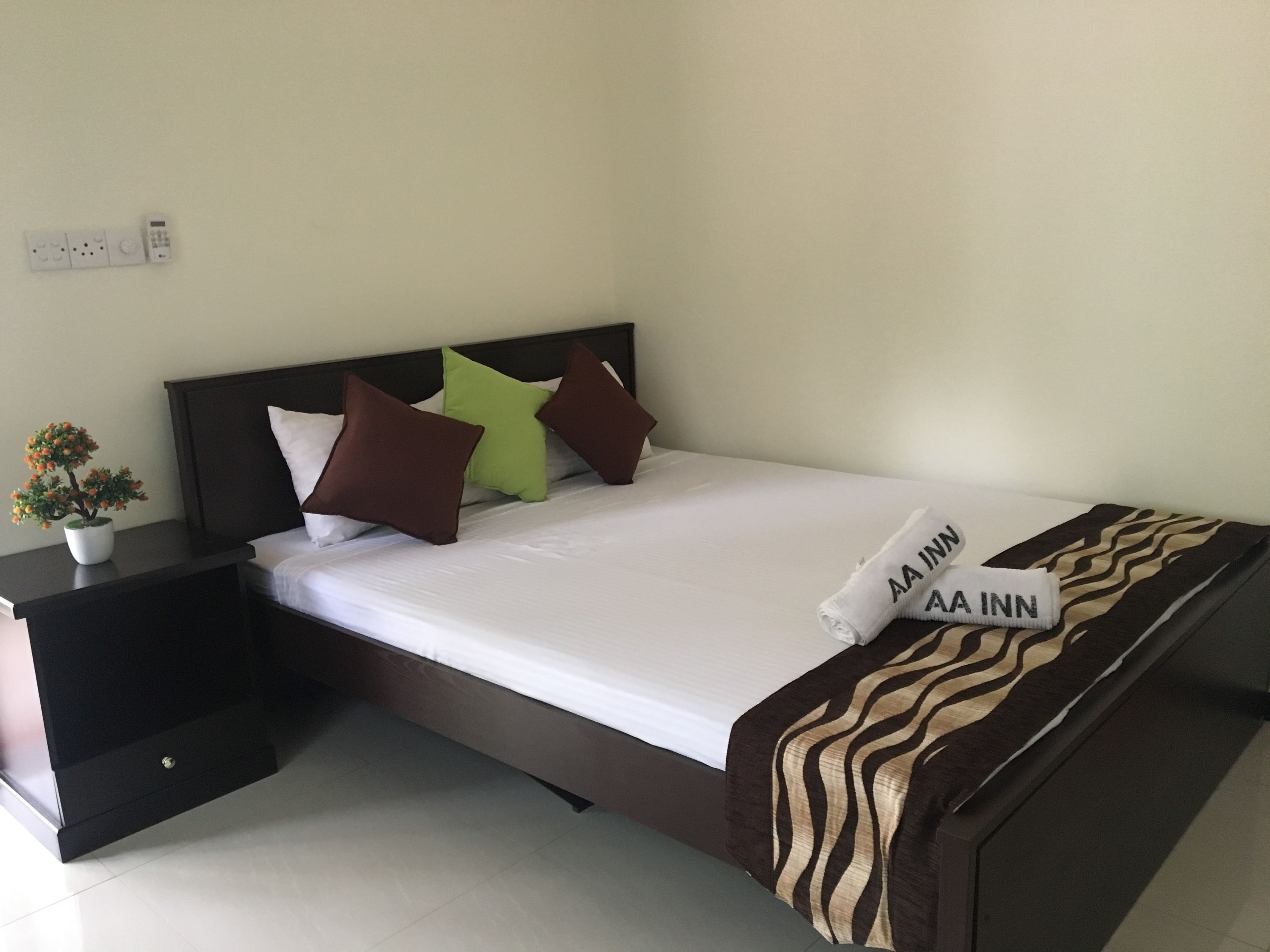 A neatly made bed with a white sheet and a brown and beige patterned blanket, a nightstand with a small potted plant, and a wall socket.