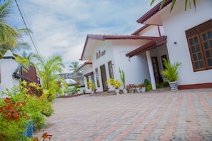 A building with a white facade and a red roof, surrounded by potted plants and palm trees. - Aainn Guest (Trincomalee)