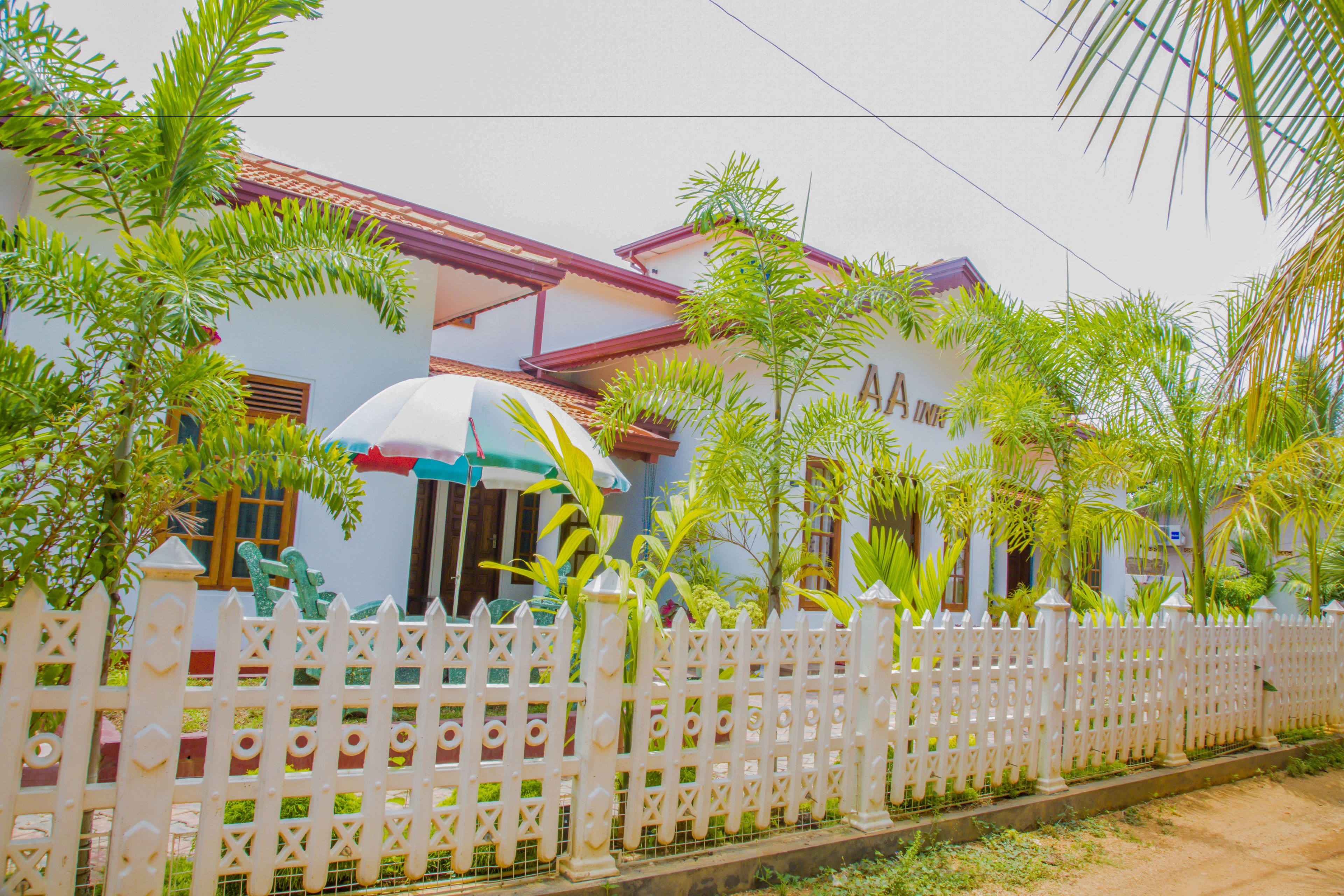 A white house with a red roof, surrounded by a white picket fence and various plants.