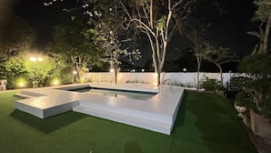 Una piscina rectangular con un deck blanco, rodeada de césped artificial y árboles.