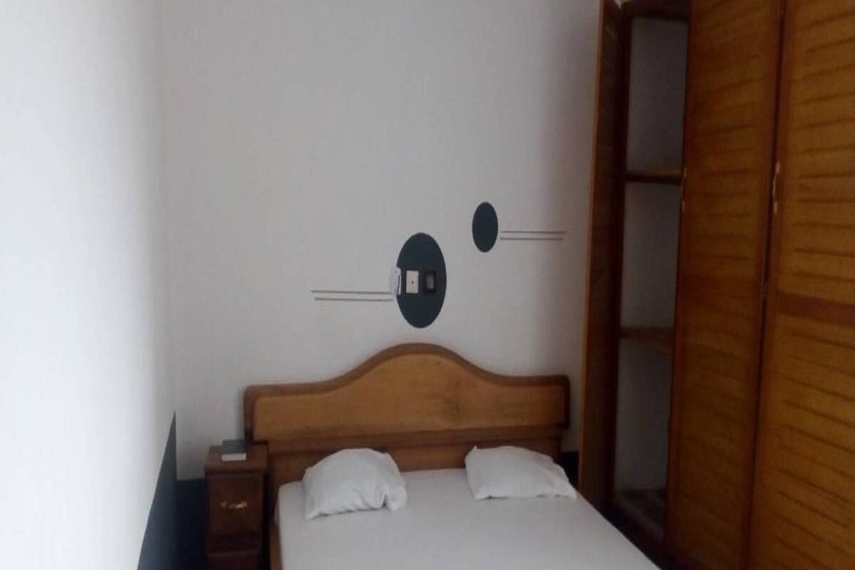 A single bed with a wooden headboard and white bedding in a room with a plain white wall and a wooden wardrobe.