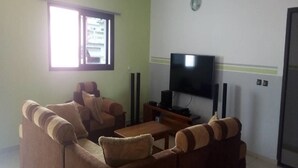 A living room with a brown sofa, a coffee table, a flat-screen TV, and a window with blinds. - Complexe Hôtelier Paris Village (Abidjan)