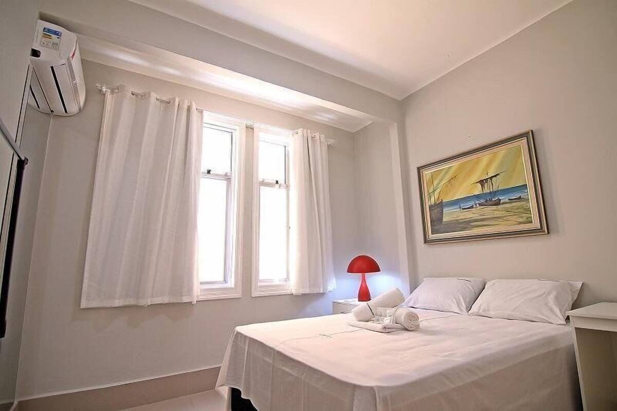Une chambre d’hôtel avec un lit, un tableau, une fenêtre avec des rideaux et une lampe rouge.
