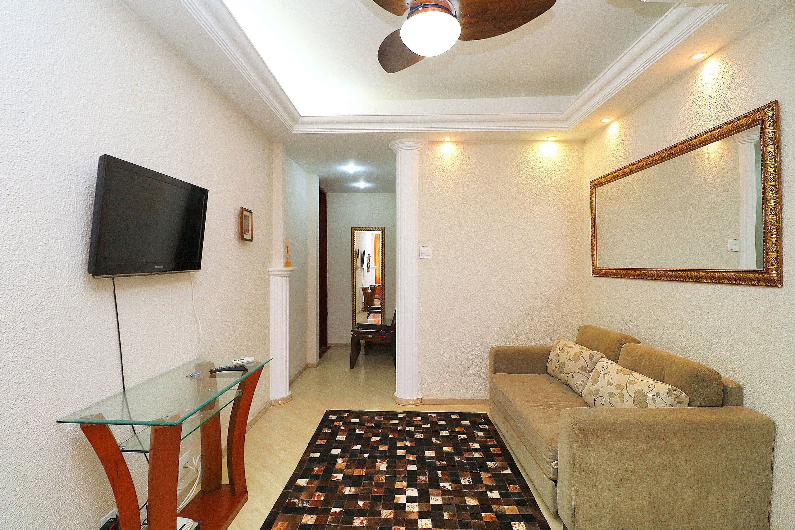 A living room with a glass coffee table, a beige sofa, a patterned rug, a television mounted on the wall, and a ceiling fan.
