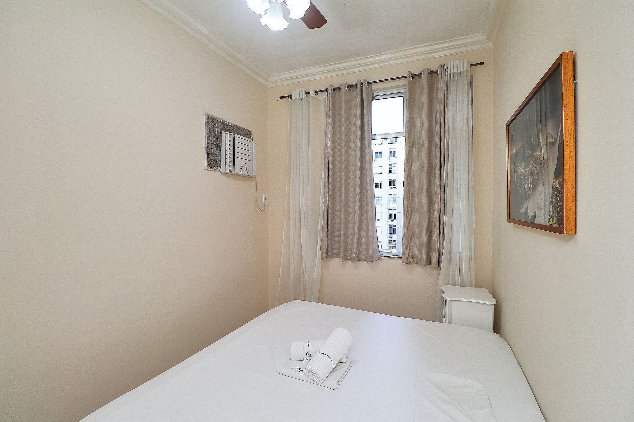 Une chambre d’hôtel avec un lit, une fenêtre avec des rideaux, un tableau au mur et une petite armoire blanche.