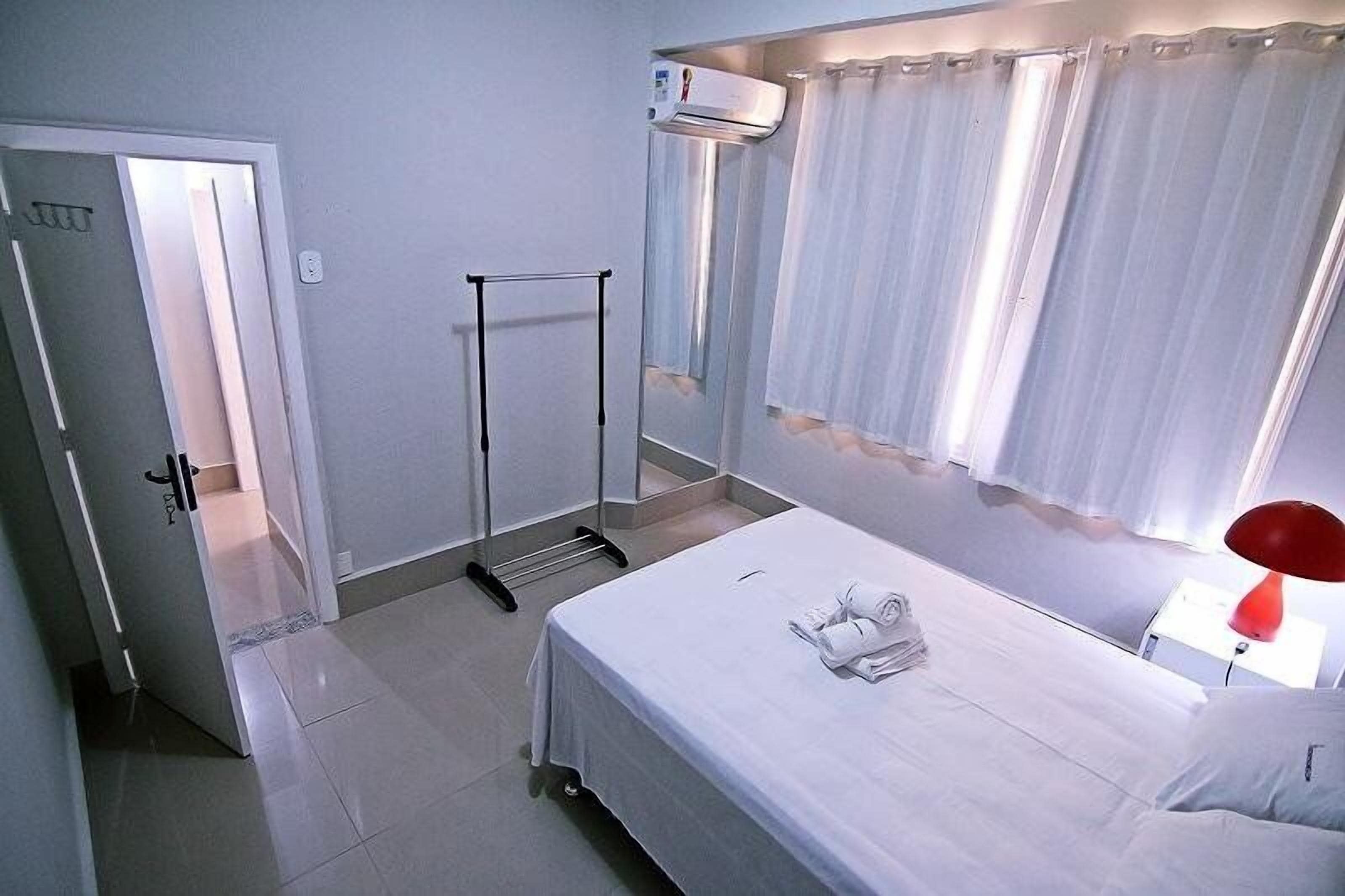 Quarto de hotel com cama, guarda-roupa, ar condicionado, janela com cortinas e uma luminária vermelha.