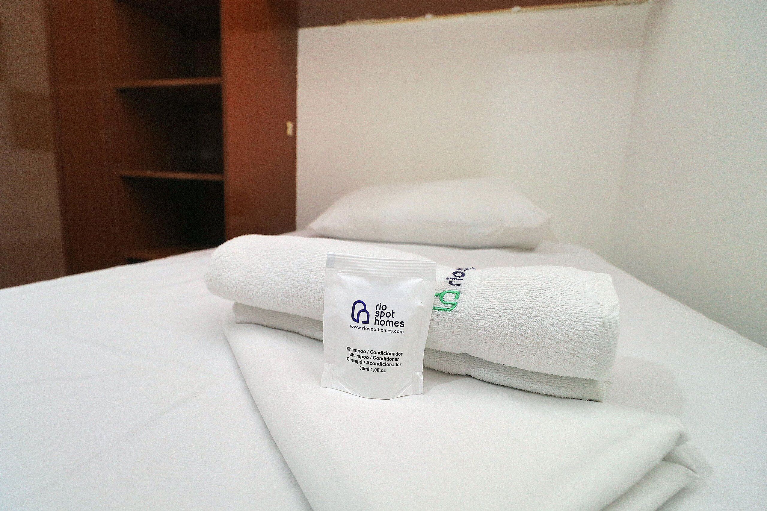 Una cama bien hecha con sábanas blancas, una toalla doblada y un pequeño paquete de champú y acondicionador.
