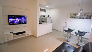A modern living room with a flat-screen TV, a sofa, and a kitchen area with a refrigerator, microwave, and bar stools. - Rio Spot Apartment D040 (Rio de Janeiro)