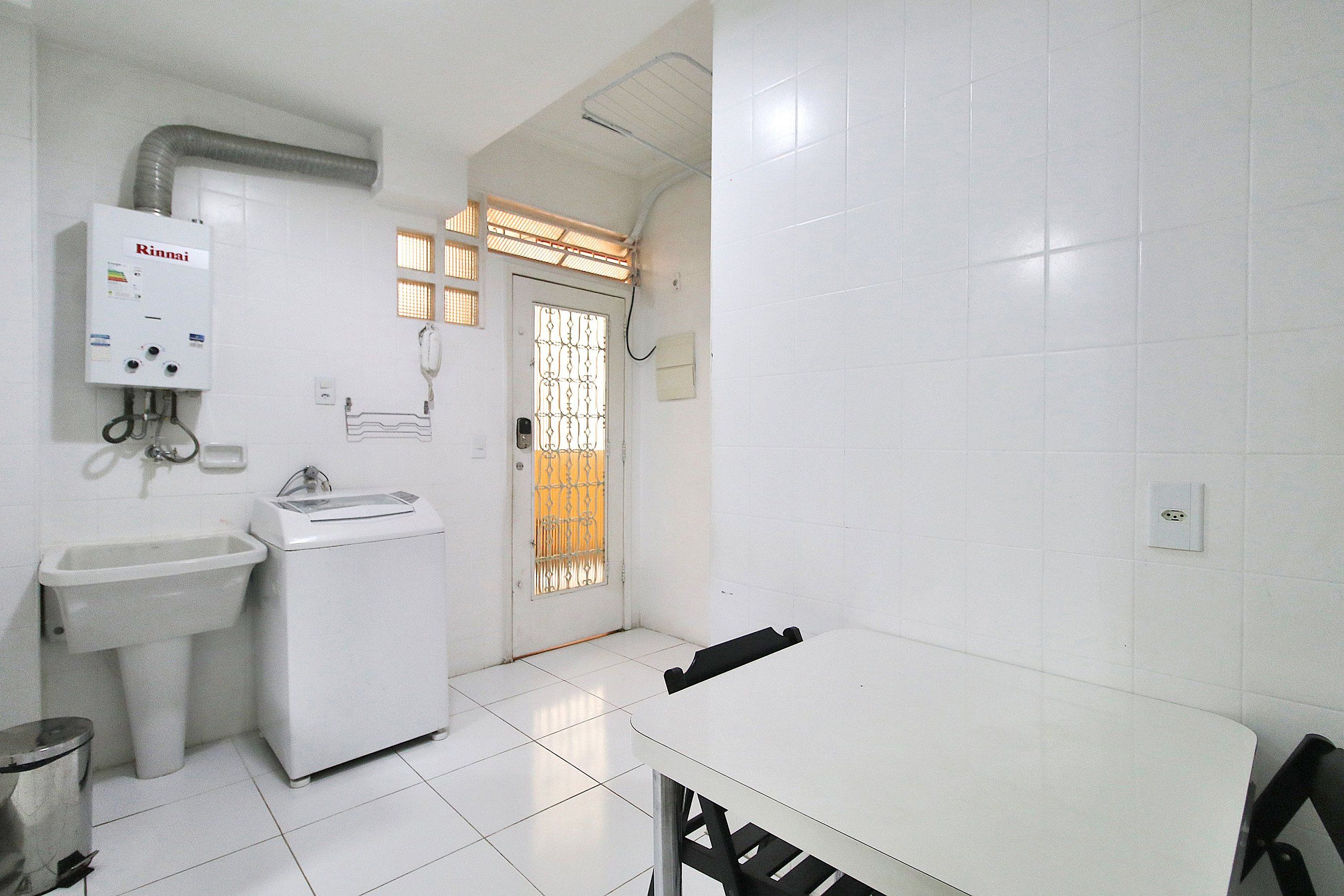 Een schone, witte badkamer met een wastafel, een wasmachine en een toilet. Er is een deur met een raam en een ventilatiesysteem erboven.