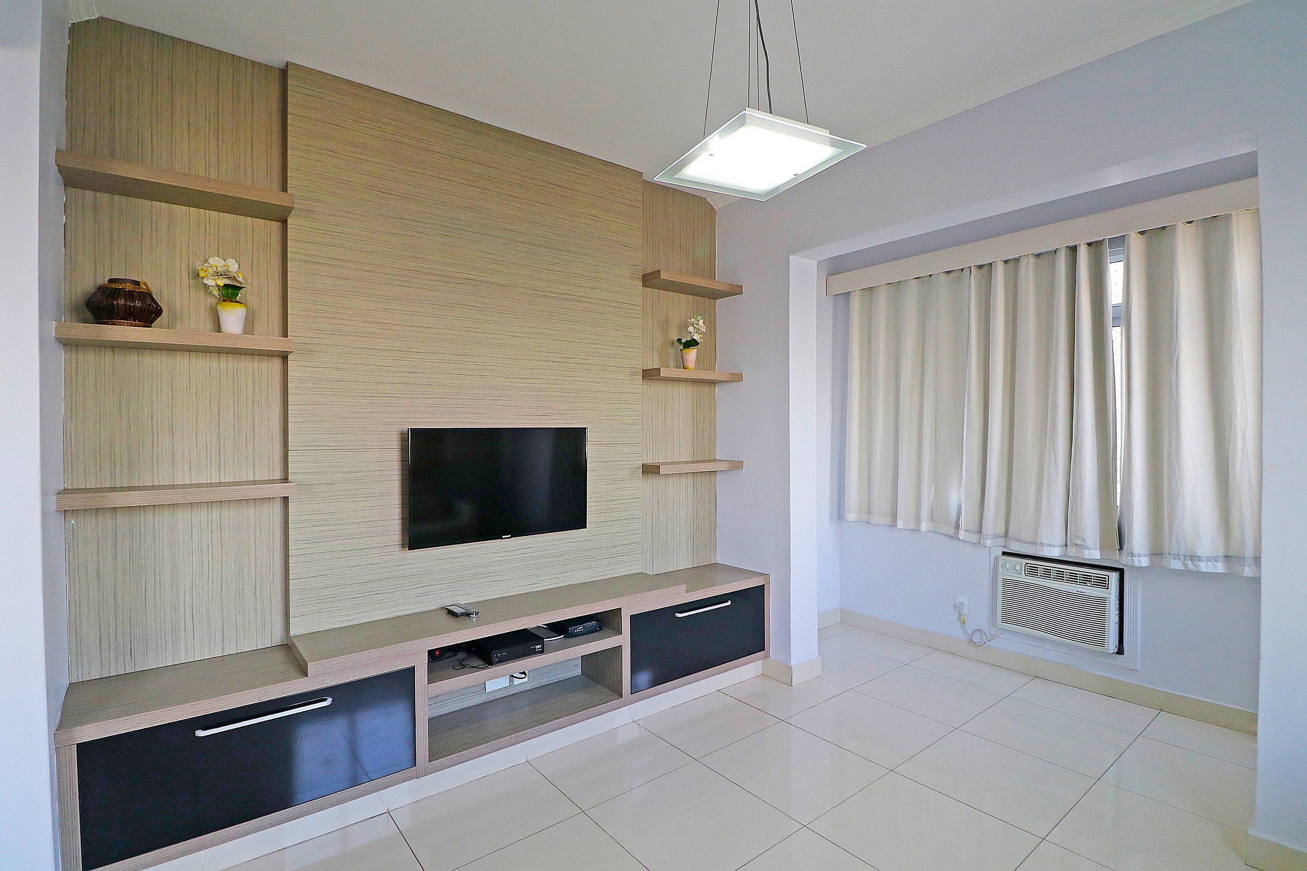 A modern living room with a flat-screen TV, wooden shelving, and a large window with curtains.