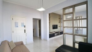 Een woonkamer met een bank, een zwarte fauteuil, een televisie op een houten plank en een groot raam met gordijnen.