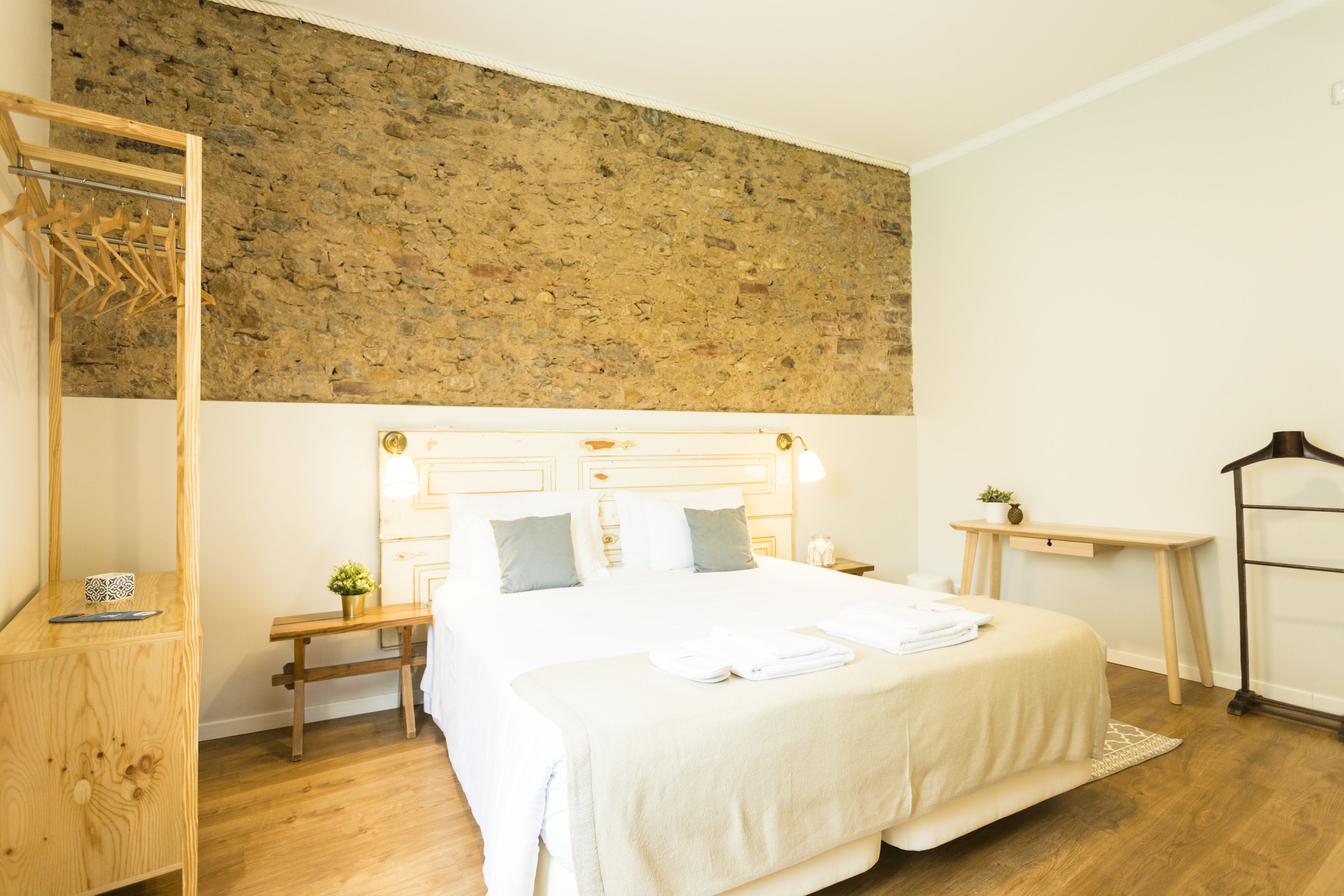 Une chambre à coucher bien aménagée, avec un grand lit, des tables de chevet, un banc en bois, un porte-vêtements et un mur en pierre.