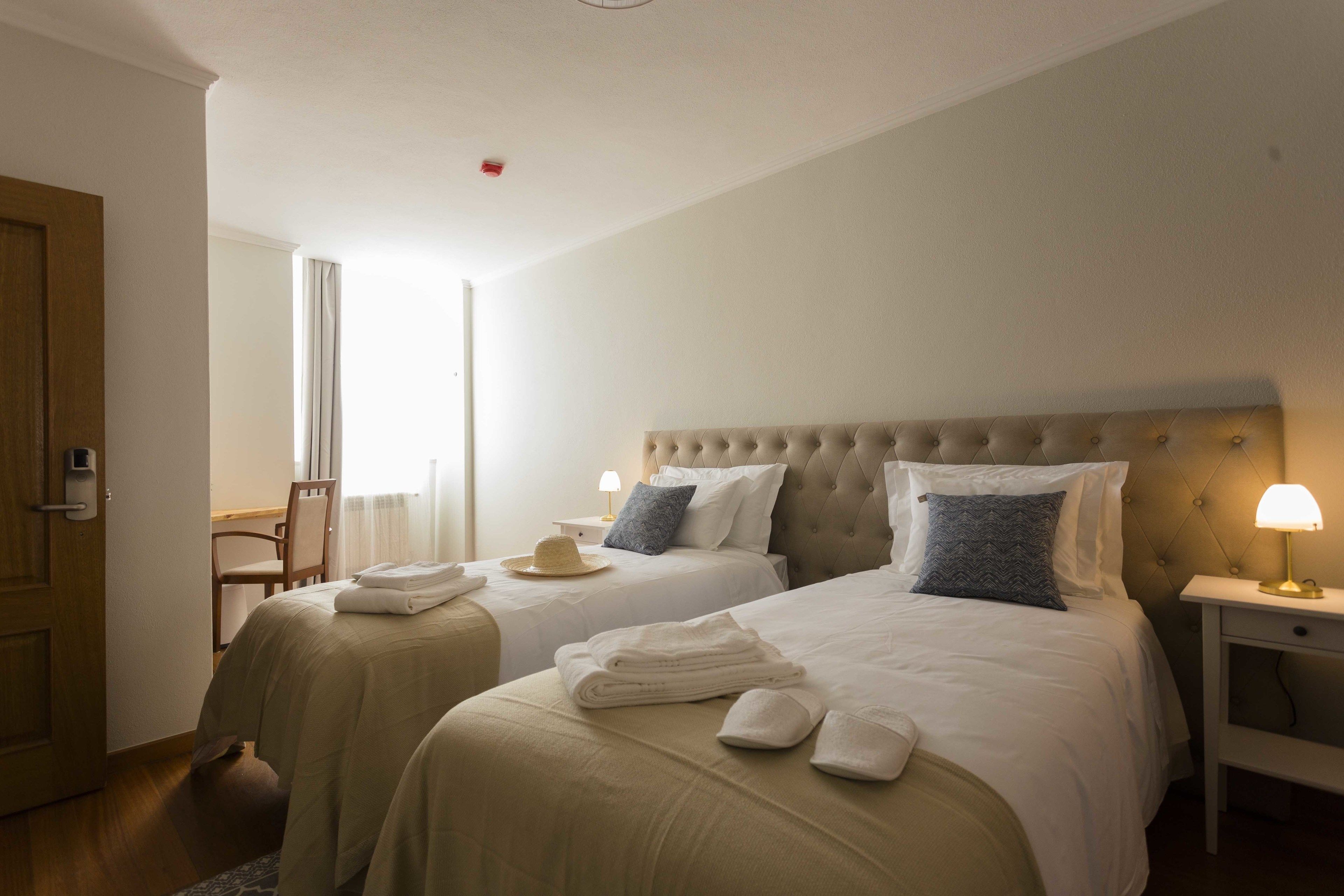 Une chambre d’hôtel avec deux lits, une table de chevet avec une lampe, une chaise et une fenêtre avec des rideaux.
