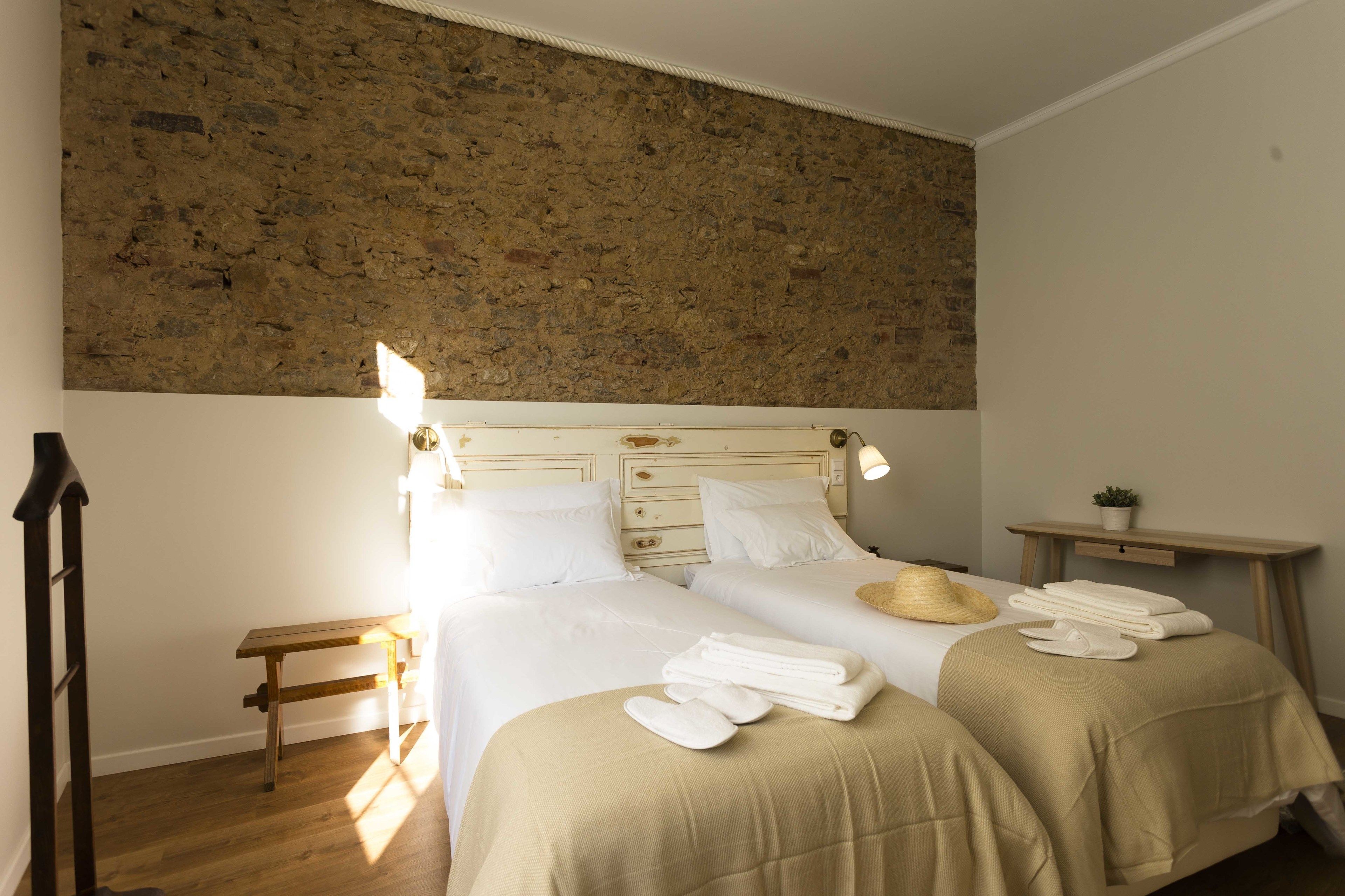 Habitación de hotel con dos camas, cabecero de madera, mesita de noche pequeña con lámpara, estante en la pared con una planta y escalera de madera apoyada contra la pared.