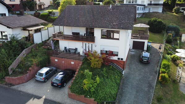 A two-story house with a sloped roof, a fenced yard, and two cars parked in the driveway.