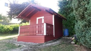 Uma pequena construção de madeira vermelha com varanda, localizada em um jardim com vegetação densa.