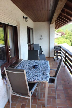 A covered patio with a table and chairs, a grill, and a view of greenery.