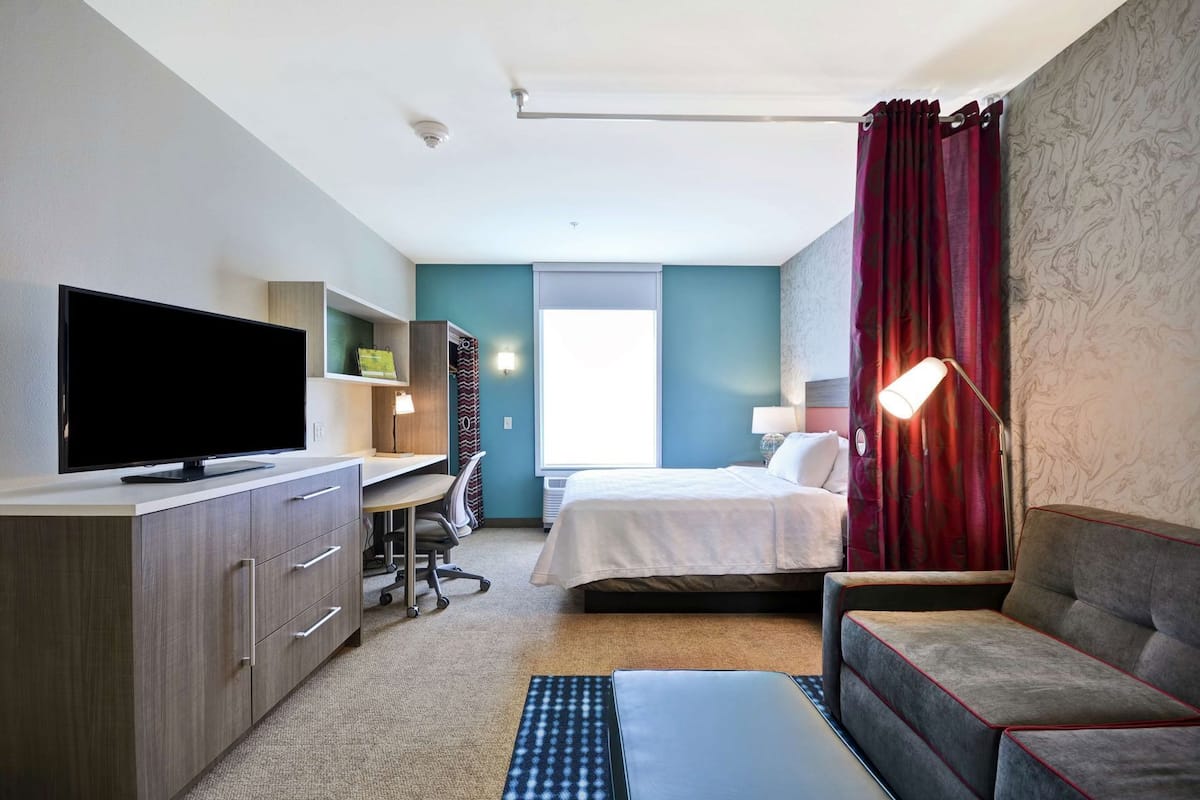 Hotelkamer met een bed, een bureau met een televisie, een bank, een lamp en een raam met gordijnen.