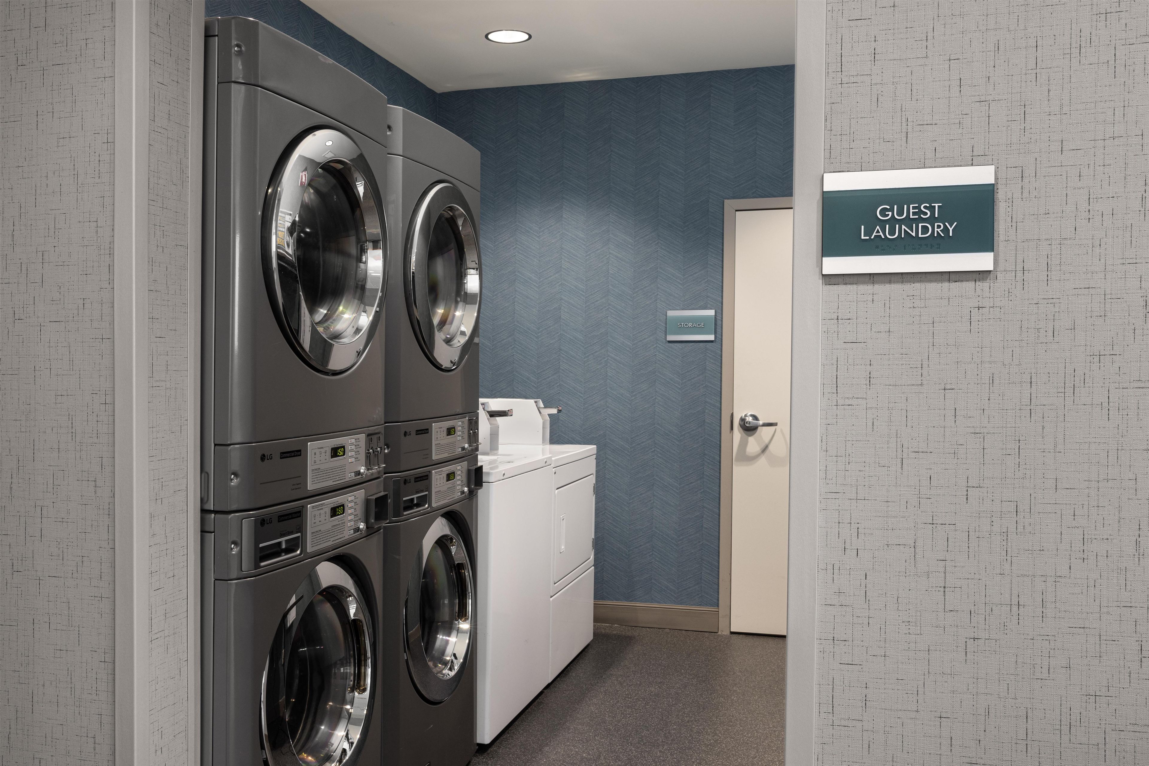 Un área de lavandería con dos lavadoras de carga frontal y una secadora, un letrero de almacenamiento y una puerta que dice 'GUEST LAUNDRY'.