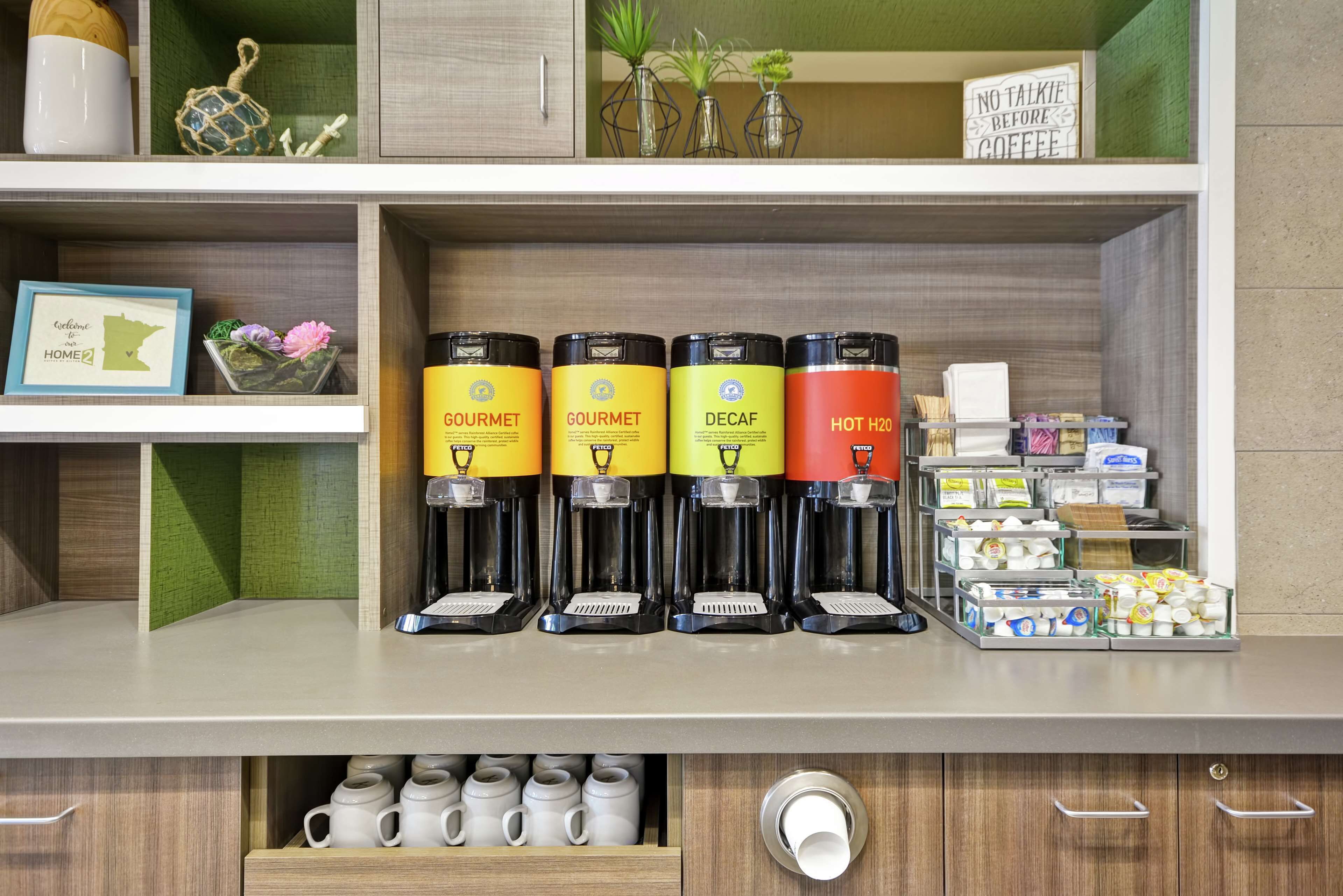 Een koffiehoek met vier koffieautomaten, gelabeld 'GOURMET', 'DECAF' en 'HEET WATER'. Ook diverse koffiekapsules en een bordje met de tekst 'GEEN KLETSEL VOOR KOFFIE'.