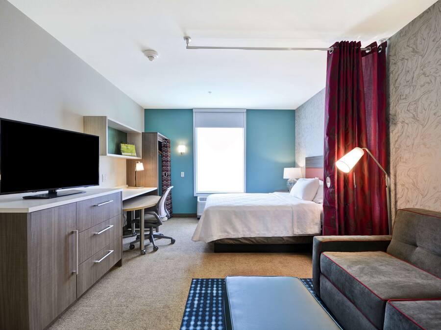 Et hotelværelse med et fladskærms-tv, et skrivebord med stol, en seng med rødt gardin og en sofa.
