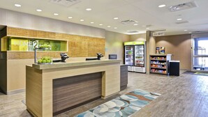 Un espace d’accueil d’hôtel comprenant un comptoir de réception en bois, un terminal d’enregistrement numérique, un réfrigérateur rempli de boissons et un distributeur automatique.