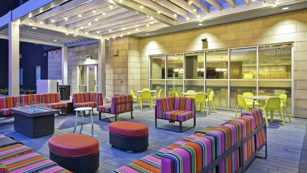Un espace extérieur aménagé en terrasse, avec des canapés et des fauteuils à rayures colorées, une table avec foyer intégré, et un restaurant avec des chaises et des tables jaunes.