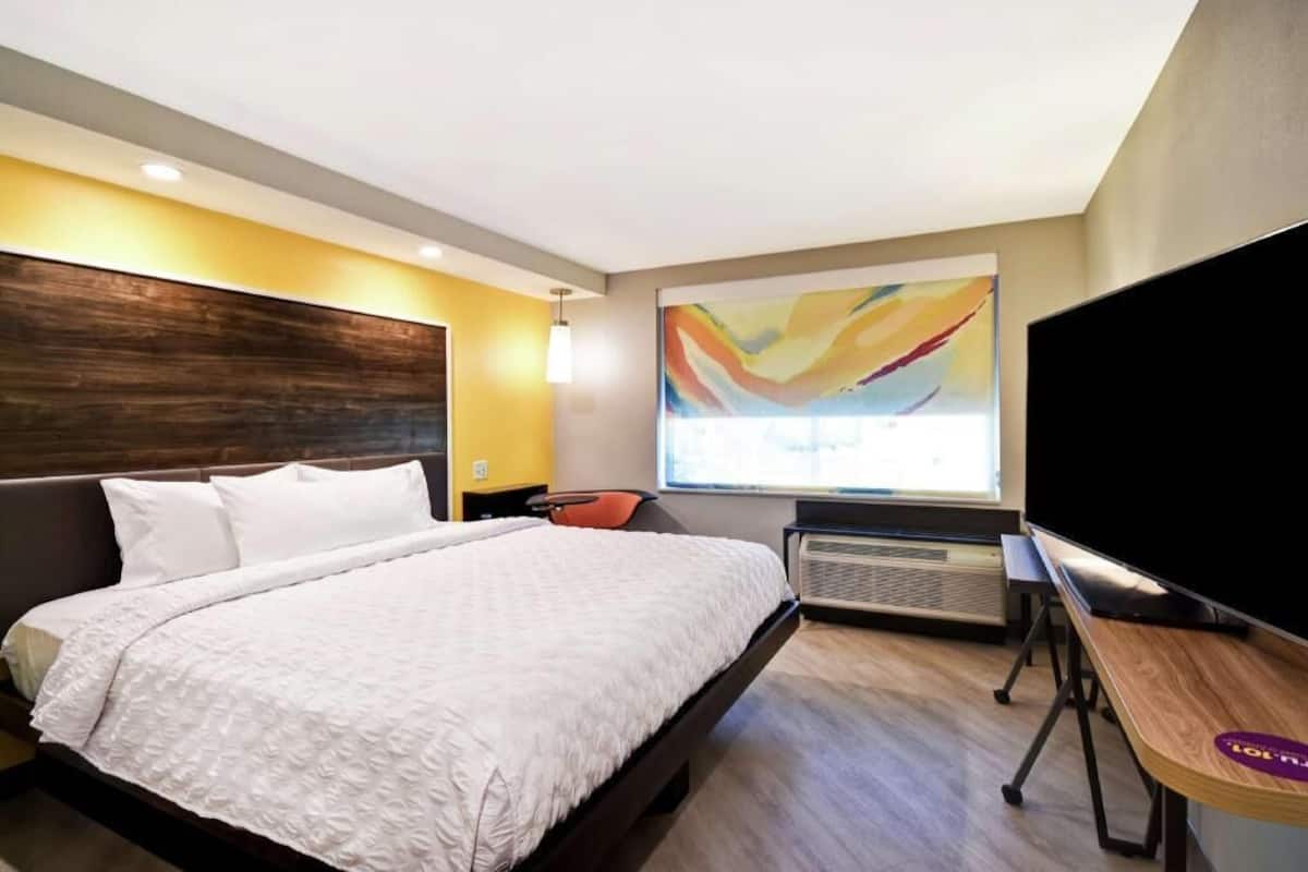 A hotel room with a bed, a desk, a chair, a TV, and a large abstract painting on the wall.