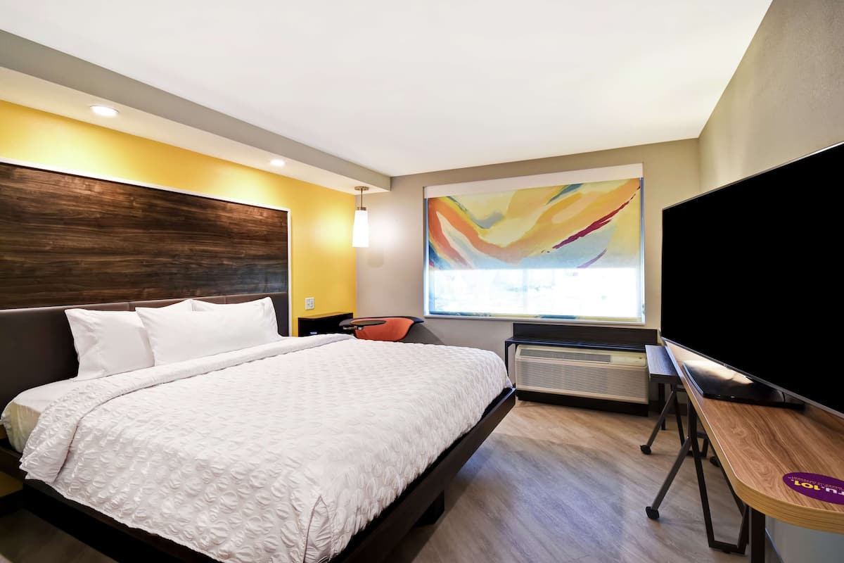 A modern hotel room with a large bed, a desk, a chair, and a large window with a colorful abstract painting.
