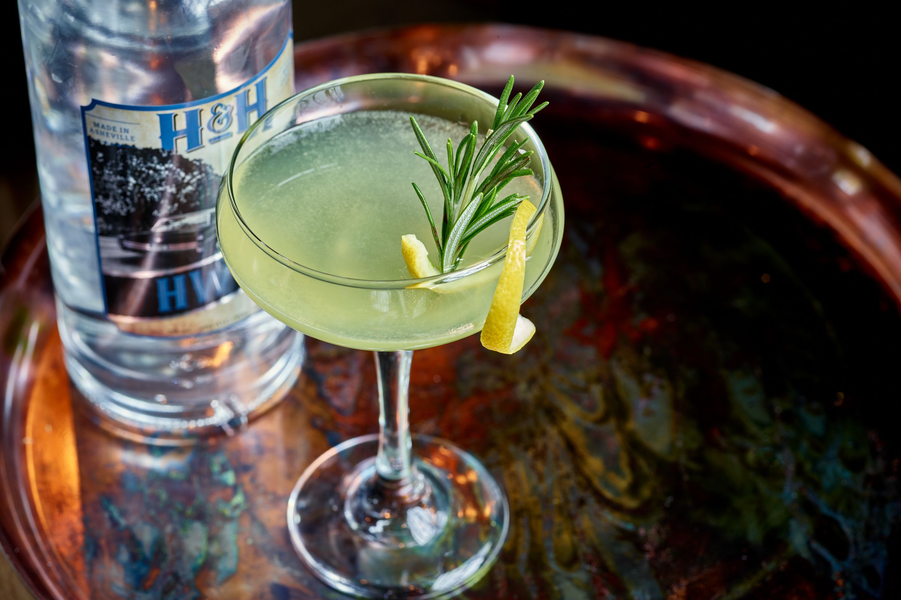 A cocktail glass with a lemon twist garnish and a sprig of rosemary, placed on a copper tray.