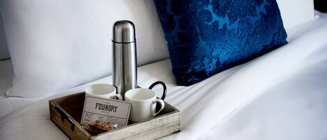 A wooden tray with a thermos, two white mugs, and a sign on a bed with white sheets and a blue patterned pillow.