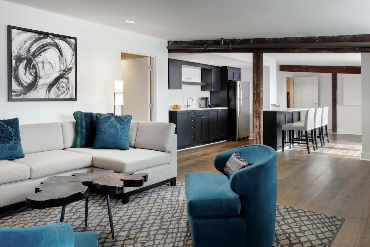 Ein modernes Wohnzimmer mit einem Ecksofa, einem blauen Sessel und einem Couchtisch. Im Hintergrund befindet sich eine Küche mit dunklen Schränken sowie ein Essbereich mit Tisch und Stühlen.