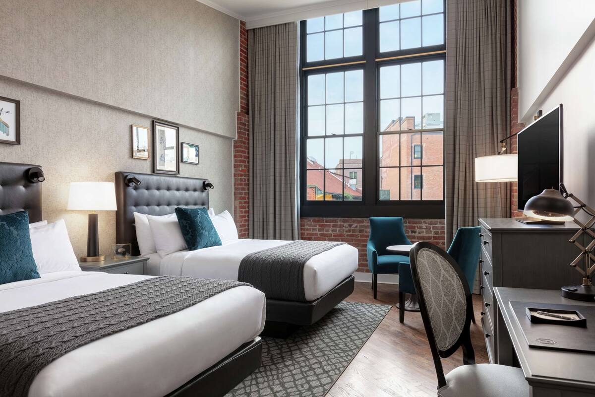 Ein Hotelzimmer mit zwei Betten, einem Schreibtisch, einem Stuhl, einem Fernseher und einem großen Fenster mit Blick auf Gebäude.