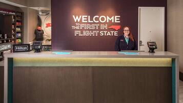 A hotel reception desk with a "Welcome to the First in Flight State" sign, a Hilton Honors badge, a phone, and a self-service kiosk.