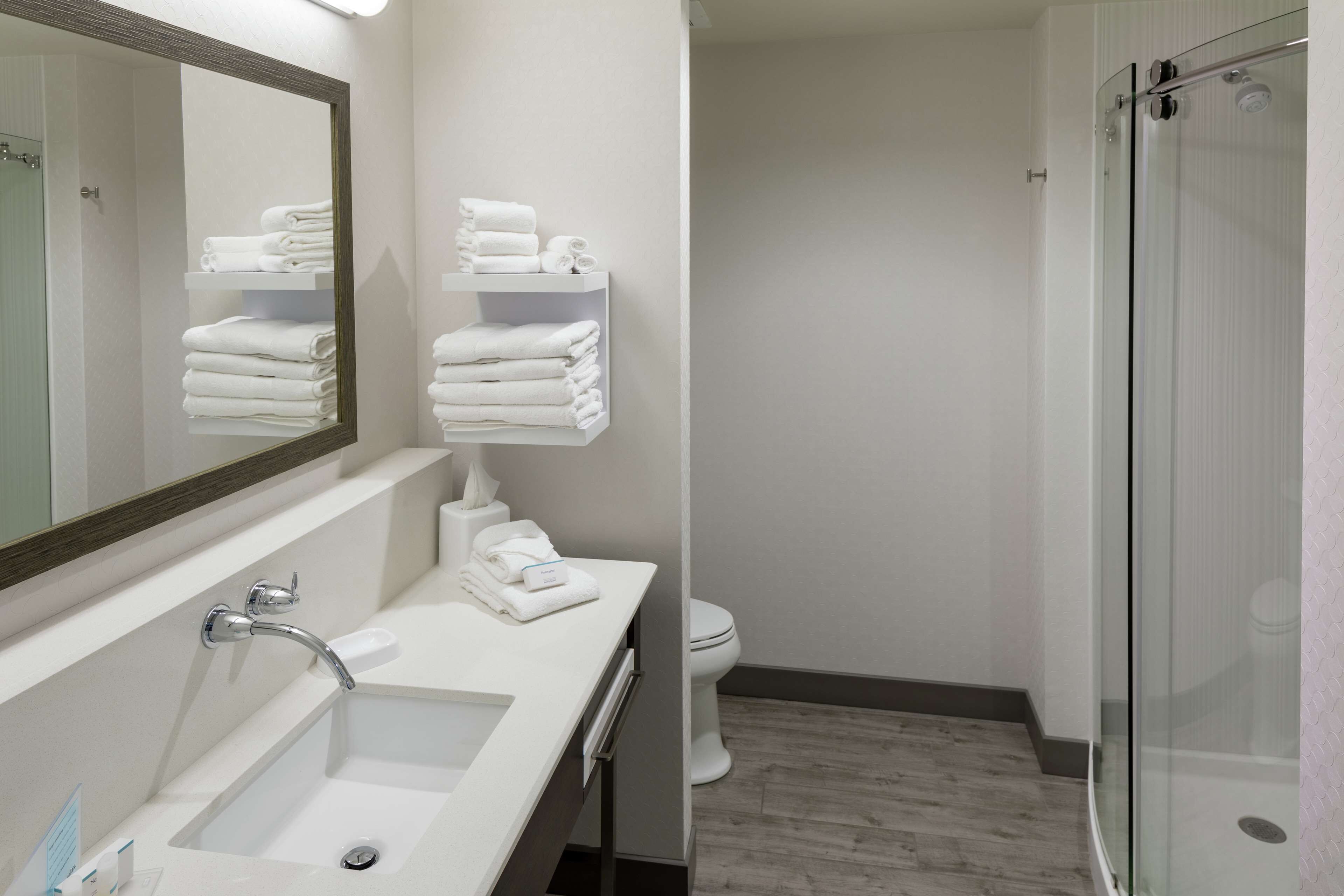 A modern bathroom with a white sink, a mirror, a glass shower enclosure, and neatly stacked towels on shelves.