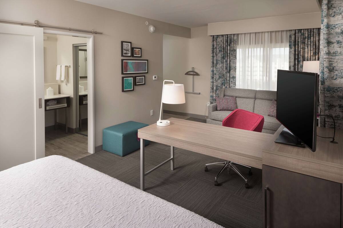 Een hotelkamer met een bed, een bureau met een lamp, een televisie, een bank en een badkamer die door een geopende deur zichtbaar is.