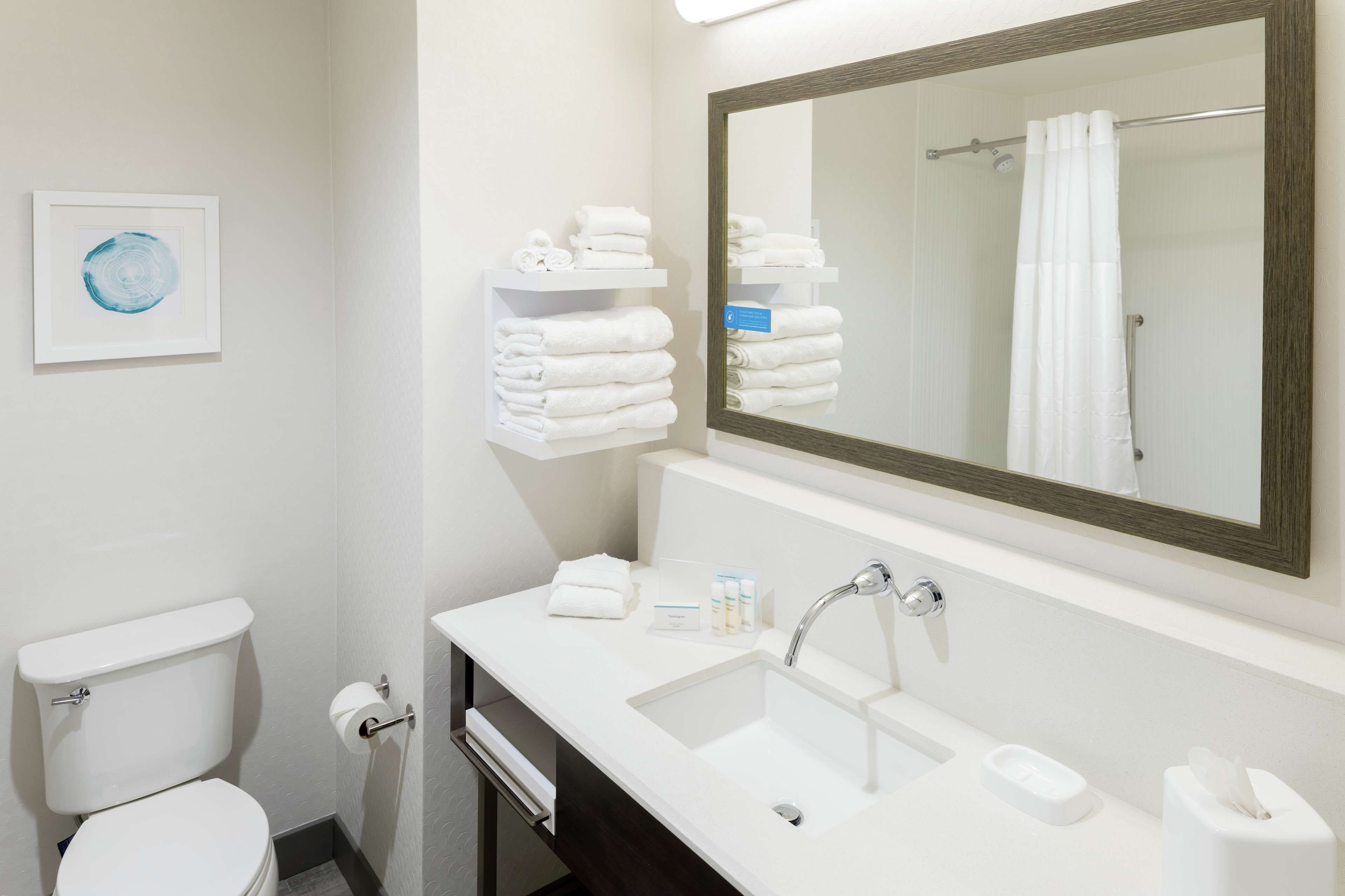 Un baño moderno con inodoro, lavamanos y espejo. Hay toallas en un estante y un cuadro en la pared.